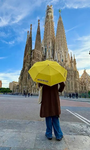 Person holding a yellow Walkative umbrella, facing the intricate spires of Sagrada Família under a blue sky in Barcelona.