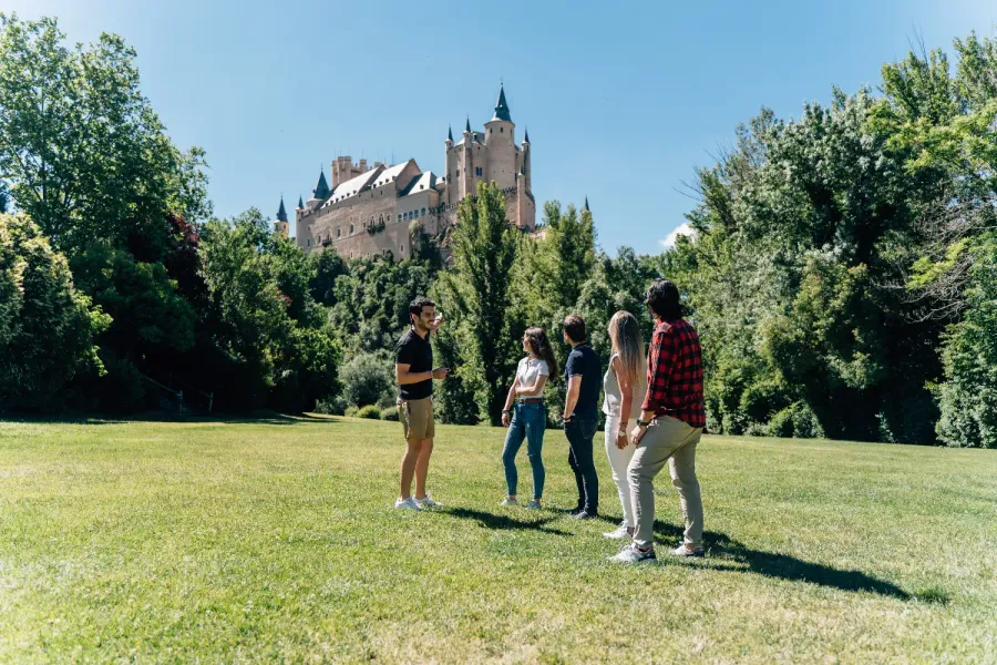 Un guía turístico habla con un grupo pequeño en un parque con el Alcázar de Segovia al fondo.