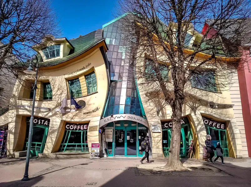 Das Krumme Haus (Krzywy Domek) in Sopot, mit geschwungener Fassade und grünem Dach, Geschäfte im Erdgeschoss, kahle Bäume.