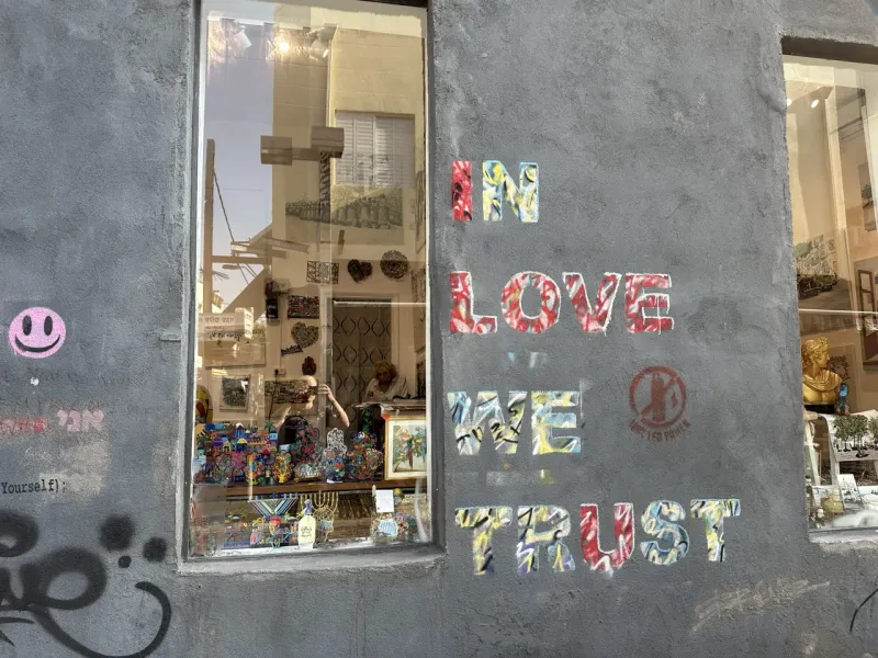 Graue Wand mit farbigem "IN LOVE WE TRUST"-Graffiti, einem rosa Smiley und einem Schaufenster, das Gebäude widerspiegelt.