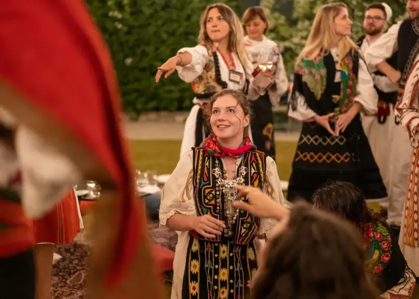 Młoda kobieta w haftowanym tradycyjnym stroju albańskim i czerwonej chuście uśmiecha się, z innymi osobami w tle.