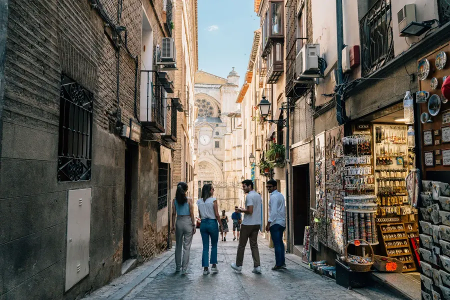 Cuatro jóvenes caminan por un callejón estrecho en Toledo, con la catedral al fondo y una tienda de recuerdos a la derecha.