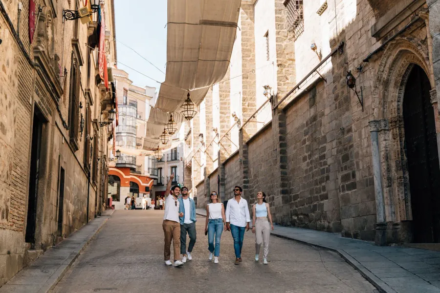 Un grupo de cinco jóvenes pasea por una estrecha calle de piedra en Toledo, a la sombra de los toldos tradicionales.