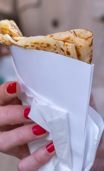 Close-up of a person's hands with red nail polish holding a sweet folded crepe wrapped in white paper.