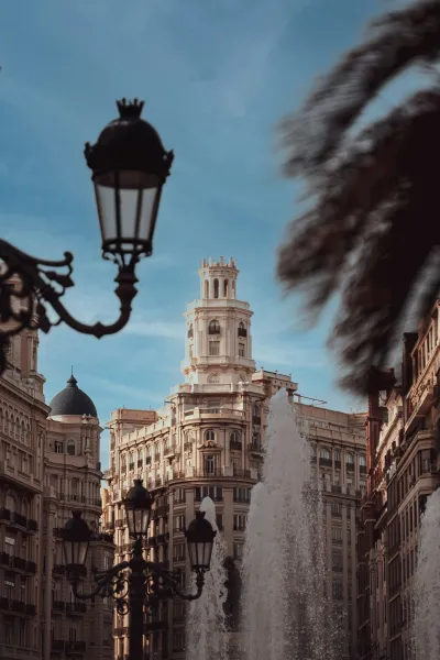 Ornate streetlamp frames a distant historic building with a tower, a fountain's spray, and palm fronds.