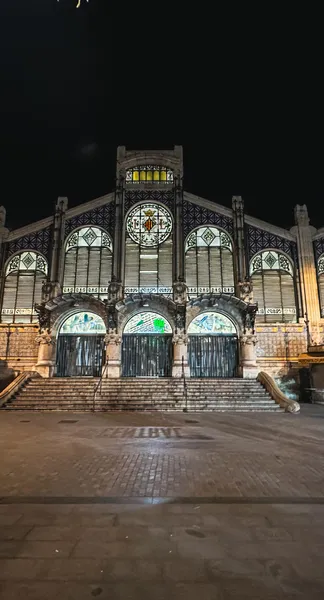 Illuminated Art Nouveau facade of Valencia Central Market at night, featuring arched entrances, steps, and intricate stain...