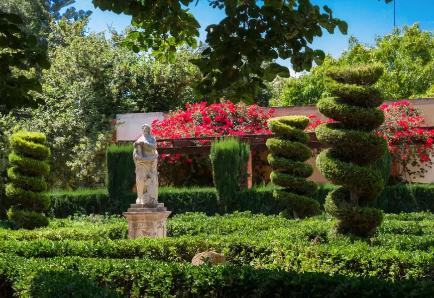 A classical statue in a green hedge garden, with spiral-shaped topiary trees and bright red flowers.