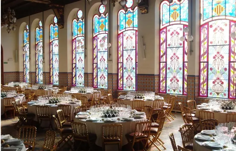 A grand hall with tables set for a banquet, illuminated by tall, colorful stained-glass windows.