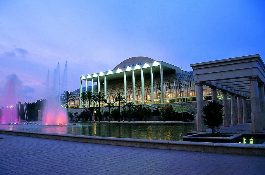 The Palau de la Música in Valencia at dusk, its glass facade lit up, with colourful fountains in the foreground.