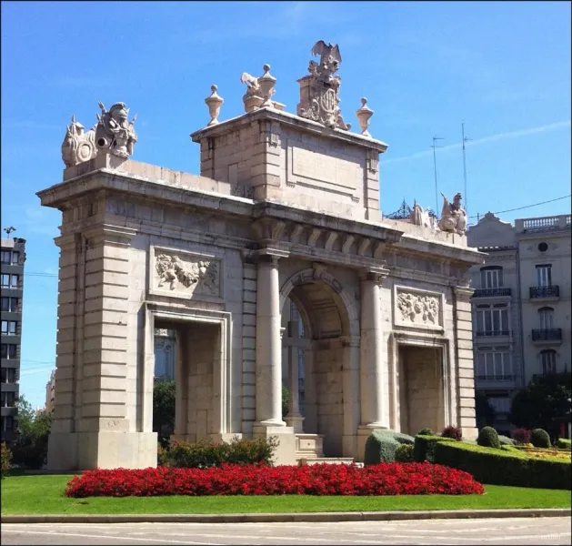 The Puerta del Mar triumphal arch in Valencia on a sunny day, with a bed of red flowers in the foreground.