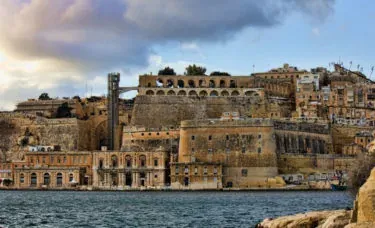 Vista de las históricas fortificaciones y edificios de piedra de La Valeta a lo largo del Gran Puerto, bajo un cielo nublado.