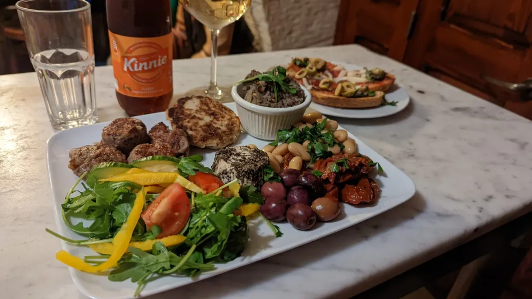 Eine Platte maltesischer Speisen mit Frikadellen, Salat, Bohnen, Oliven und Käse, dazu eine Kinnie-Flasche und Weinglas au...