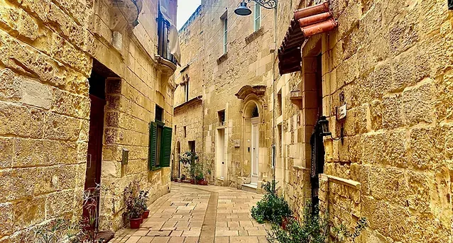 Una calle estrecha de piedra en un casco antiguo, flanqueada por edificios altos con balcones, persianas verdes y farolas.