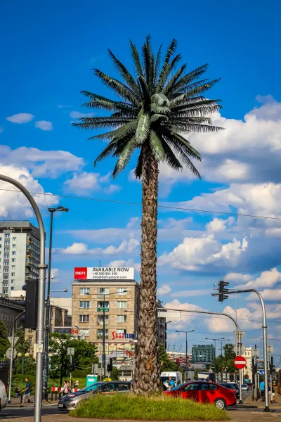 Hohe künstliche Palme auf einer Grüninsel an einer Stadtkreuzung, mit Gebäuden, Verkehr und Fußgängern unter blauem Himmel.