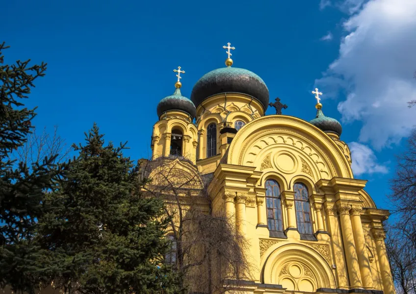Yellow Orthodox church with dark domes and golden crosses against a blue sky, surrounded by green trees.