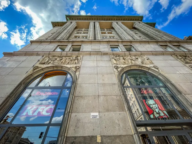 Untersicht eines grauen Steingebäudes mit klassischen Säulen und Reliefschnitzereien. Große Bogenfenster zeigen Reflexione...