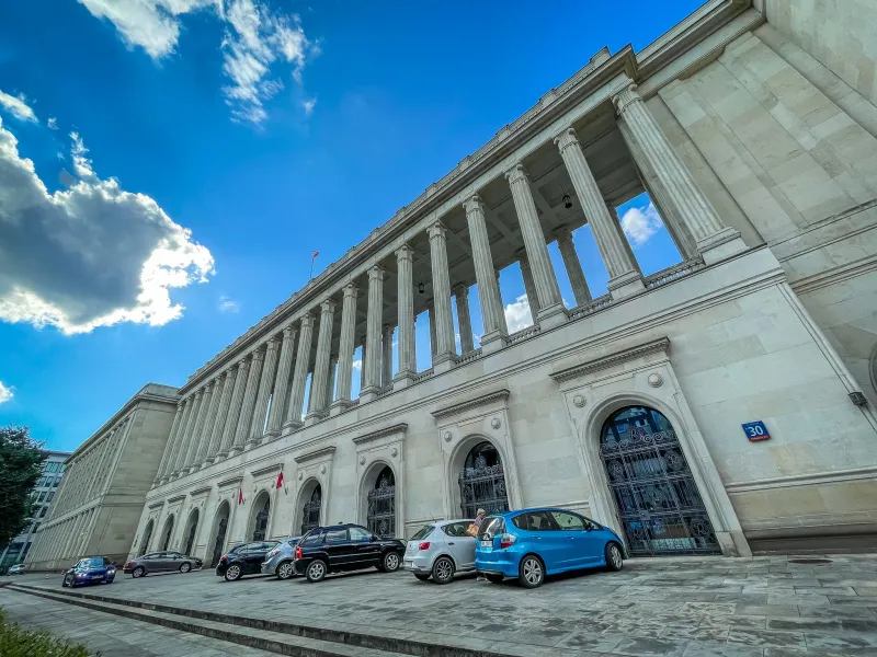 Large, imposing building with a long colonnade and parked cars under a bright blue sky in Warsaw.