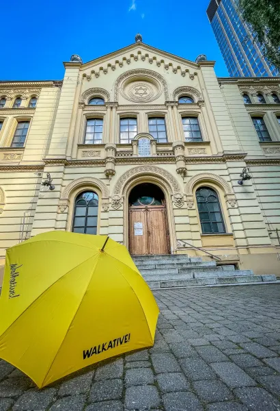 Front facade of the ornate Nożyk Synagogue in Warsaw, a yellow Walkative umbrella on stone steps in the foreground.