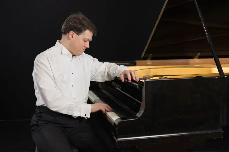 A male pianist in a white tuxedo shirt and black trousers plays a black grand piano against a dark background.