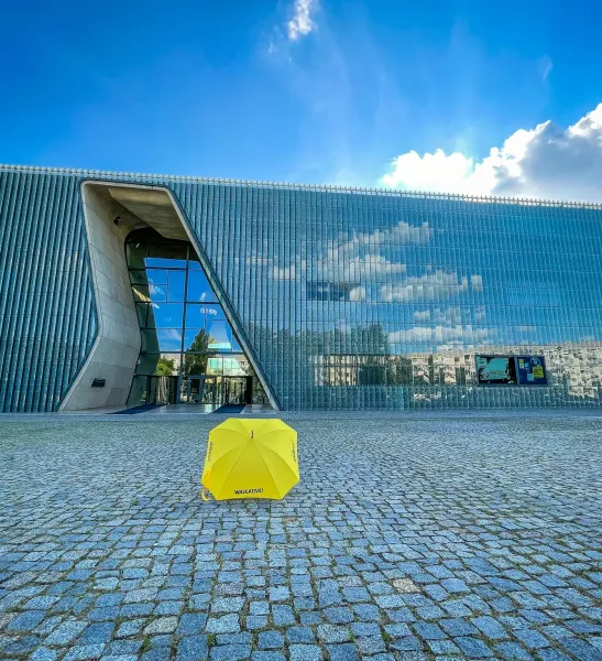 Paraguas amarillo Walkative en plaza adoquinada frente al moderno edificio de cristal del Museo POLIN en Varsovia.