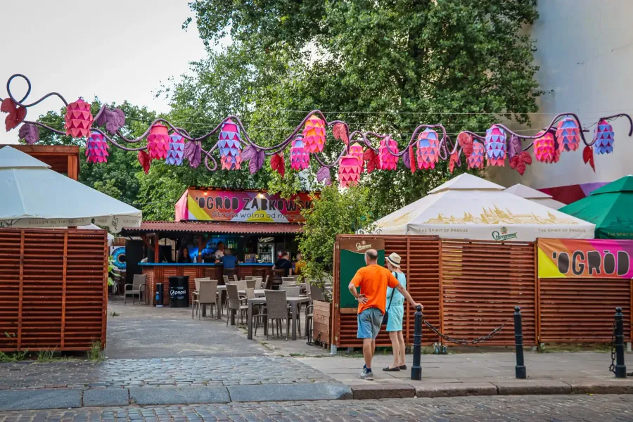 Outdoor bar area under colorful hop-shaped decorations, with tables, chairs, and two people standing on a cobblestone street.