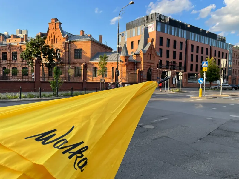 A yellow Walkative umbrella in the foreground, with historical brick buildings, a modern building, and a street in Warsaw'...