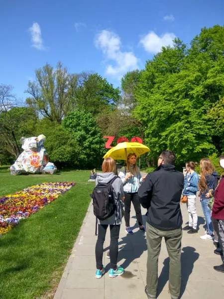 Tour guide with yellow umbrella speaking to a group on a paved path, next to a flower bed and green trees. A ZOO sign and ...