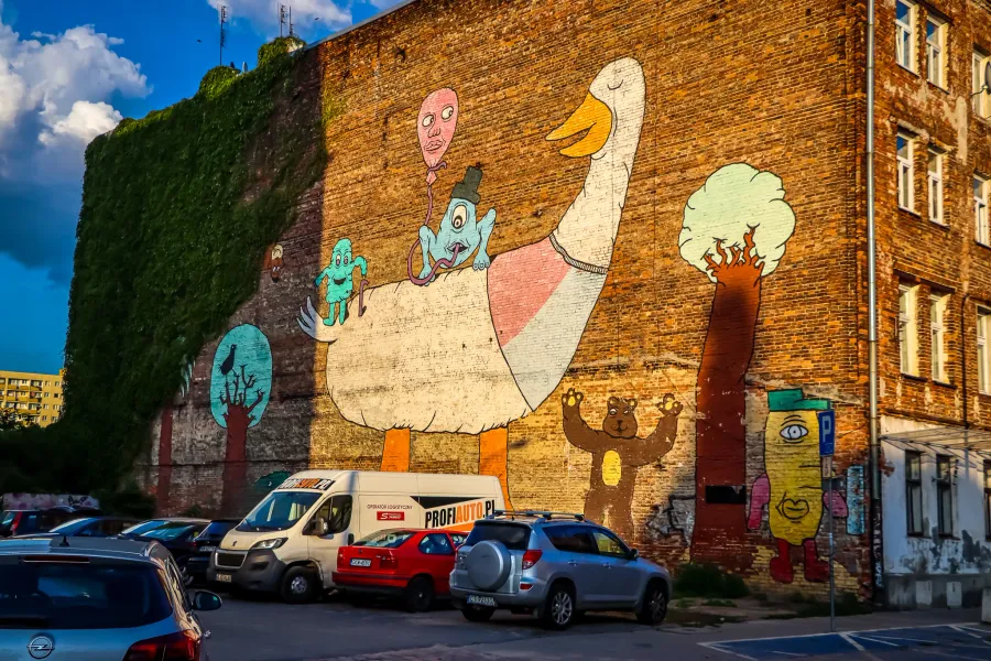 Colorful mural on a brick building in Warsaw, showing a large goose, whimsical figures, and a bear, with parked cars below.