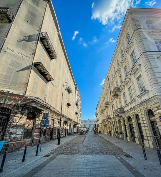Calle adoquinada entre un edificio andamiado con 'warimpex' y un edificio histórico ornamentado con balcones bajo cielo azul.