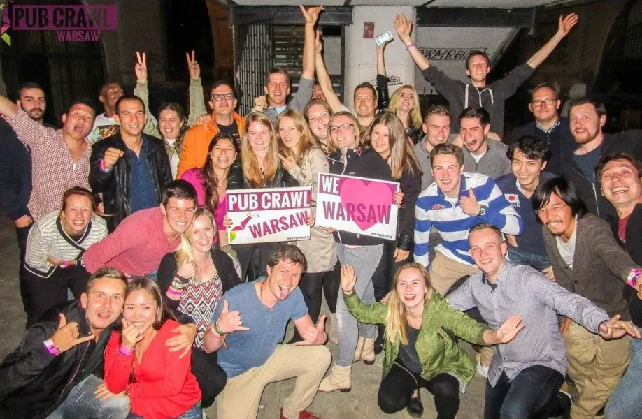 Eine große Gruppe junger Erwachsener lächelt und posiert mit „Pub Crawl Warsaw“- und „Wir lieben Warschau“-Schildern in st...