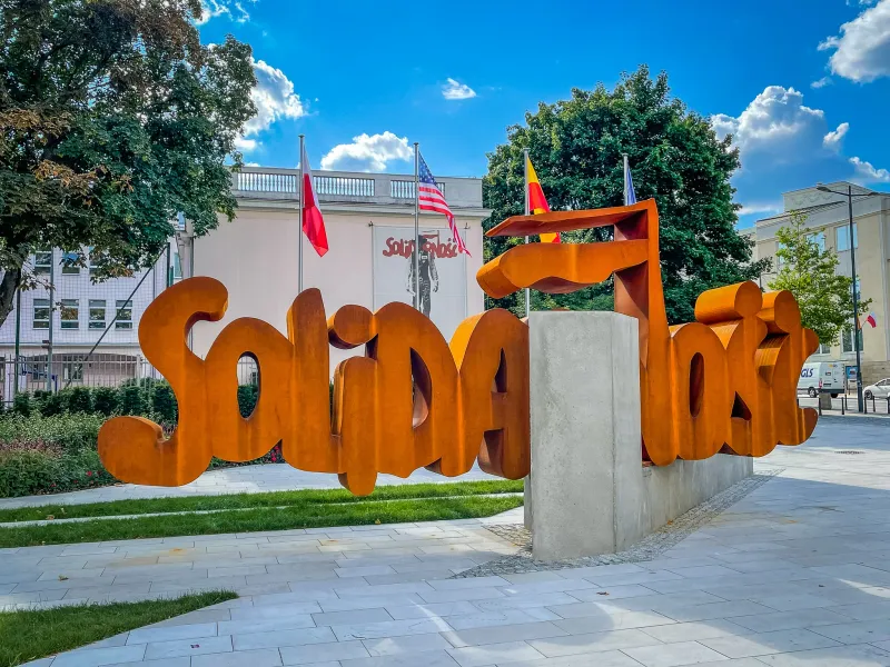 Rostfarbene Skulptur mit 'Solidarność' in Warschau. Polnische und US-Flaggen, Bäume und ein weißes Gebäude im Hintergrund.