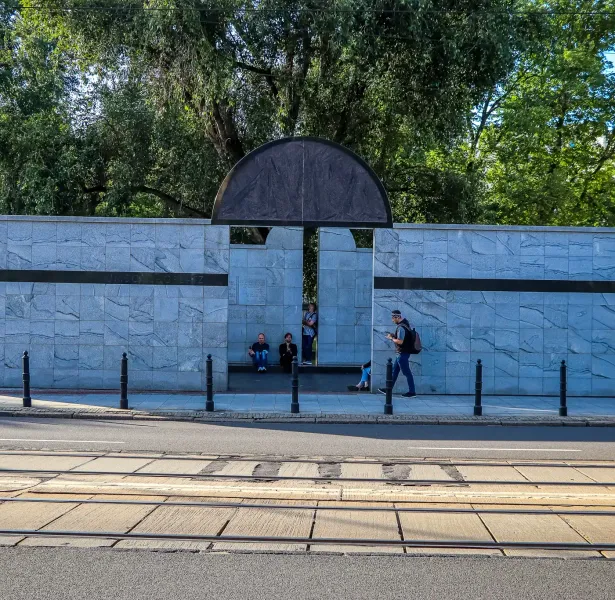 Muro conmemorativo de mármol gris con arco semicircular oscuro, personas de pie y sentadas, vías de tranvía y calle en pri...
