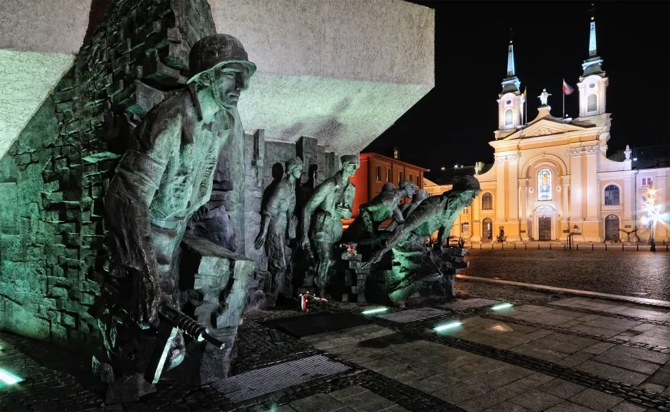 Bronzemonument mit Soldaten des Warschauer Aufstands, nachts grün beleuchtet, im Hintergrund eine hell erleuchtete Kirche.