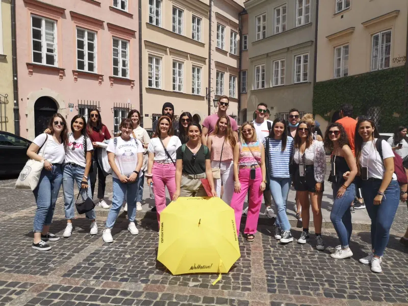 Un grupo de jóvenes posa en una calle empedrada con un paraguas amarillo de Walkative. Edificios históricos coloridos al f...