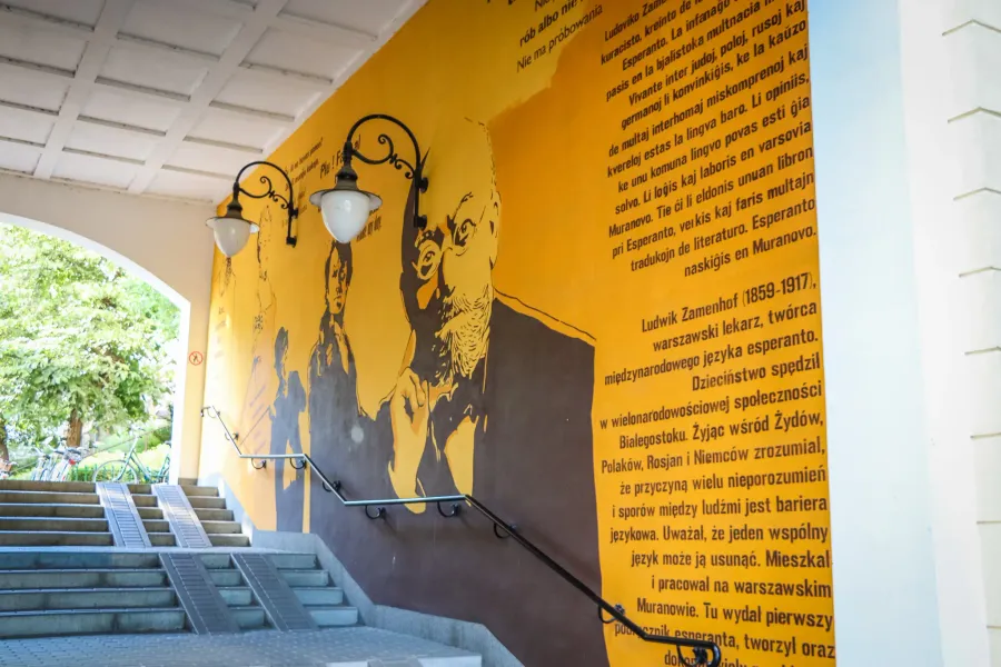 Mural amarillo de Ludwik Zamenhof con texto, junto a escaleras de cemento con pasamanos y farolas.
