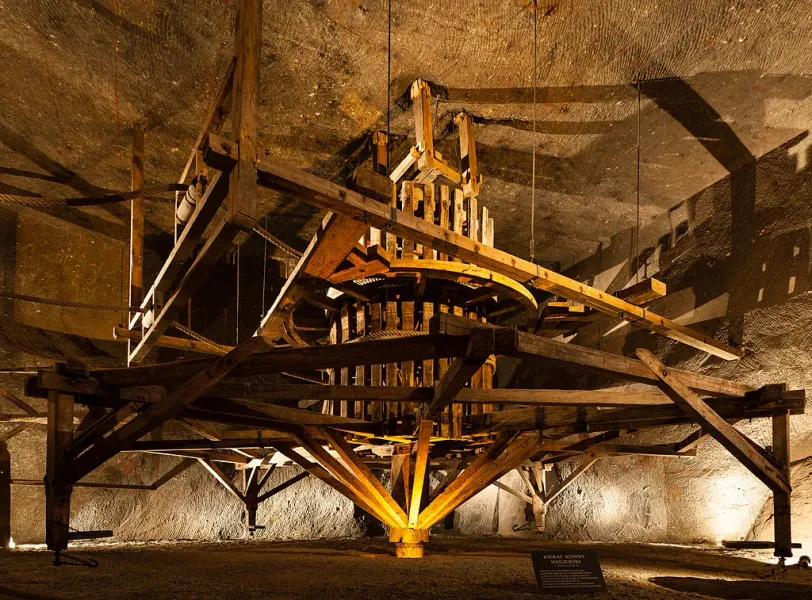 A large, historical wooden horse-powered hoisting machine inside the Wieliczka Salt Mine, with salt rock walls behind it.