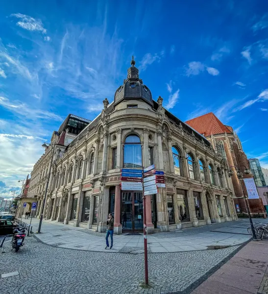 Ozdobny zabytkowy budynek z kopulastą wieżą narożną i dużymi oknami przy brukowanej ulicy we Wrocławiu. Widoczny drogowskaz.