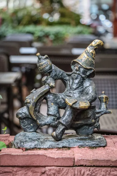 Bronze statue of a Wroclaw dwarf riding a scooter. The dwarf wears a pointy hat and holds a bottle of Chianti. A moka pot ...