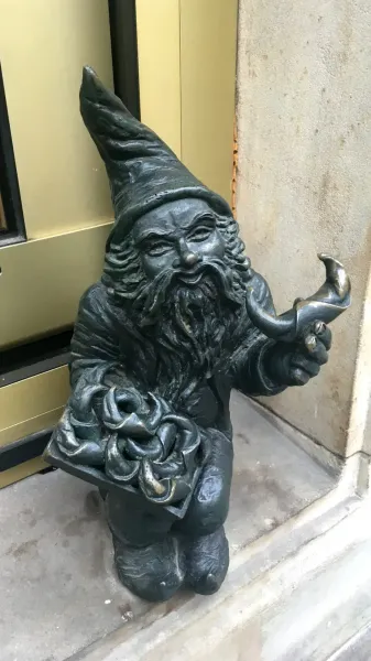 Bronze dwarf statue with a pointed hat and long beard, holding a tray with knots and a horn-shaped object, next to a build...