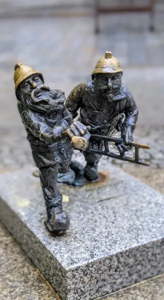 Dwie ciemne, brązowe figurki krasnali z Wrocławia w złotych hełmach, jeden z brodą trzymający kilof, drugi drabinę.