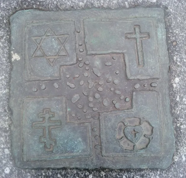 Brązowa tablica w granicie z czterema symbolami religijnymi: Gwiazda Dawida, krzyż łaciński, krzyż prawosławny i róża Lutra.