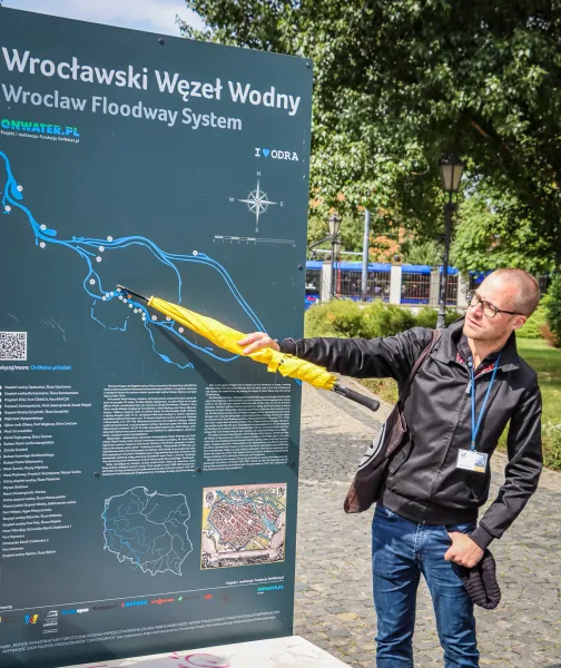 Mann zeigt mit gelbem Regenschirm auf eine Informationstafel mit Karte des Breslauer Wasserstraßensystems an einem sonnige...