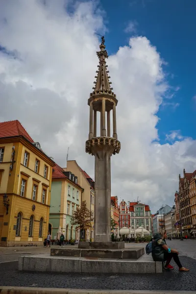 Wysoki kamienny pręgierz z ozdobną iglicą i figurą na szczycie, otoczony kolorowymi zabytkowymi budynkami na rynku.