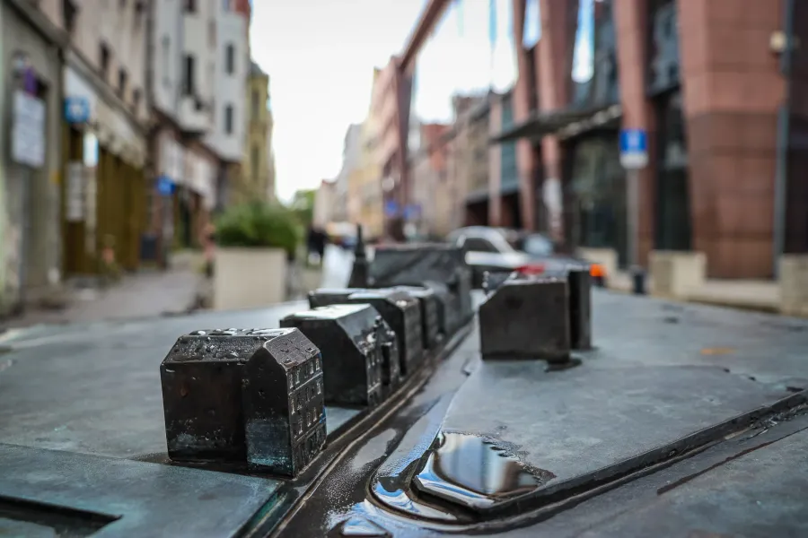 Close-up of a dark metal miniature model of city buildings with water puddles, blurring into a real street scene in the ba...