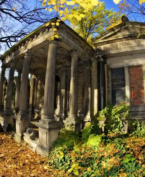 Stone mausoleum with classical columns, intricate stonework, and green ivy, surrounded by fallen yellow autumn leaves unde...