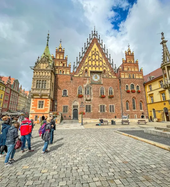Wrocławski Ratusz, duży gotycki budynek z cegły z wysoką wieżą zegarową, stoi na brukowanym Rynku z grupą ludzi.