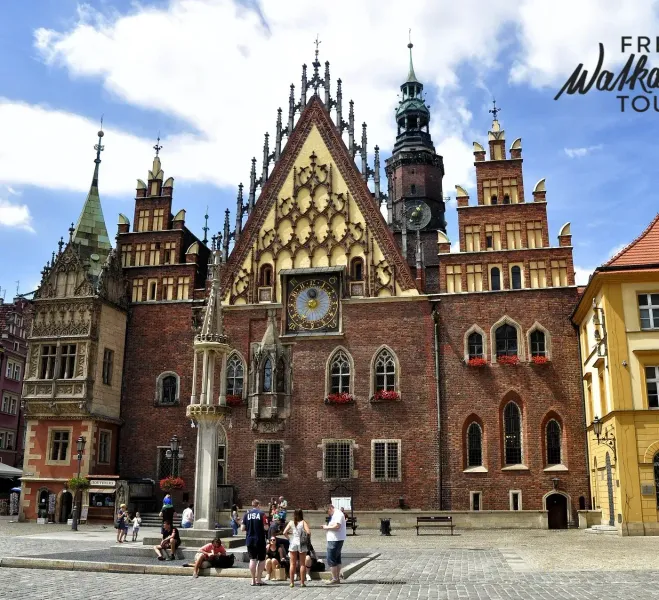 Gotycki ceglany Ratusz we Wrocławiu z dużym zegarem, ozdobnymi szczytami i wysoką wieżą. Ludzie gromadzą się na brukowanym...