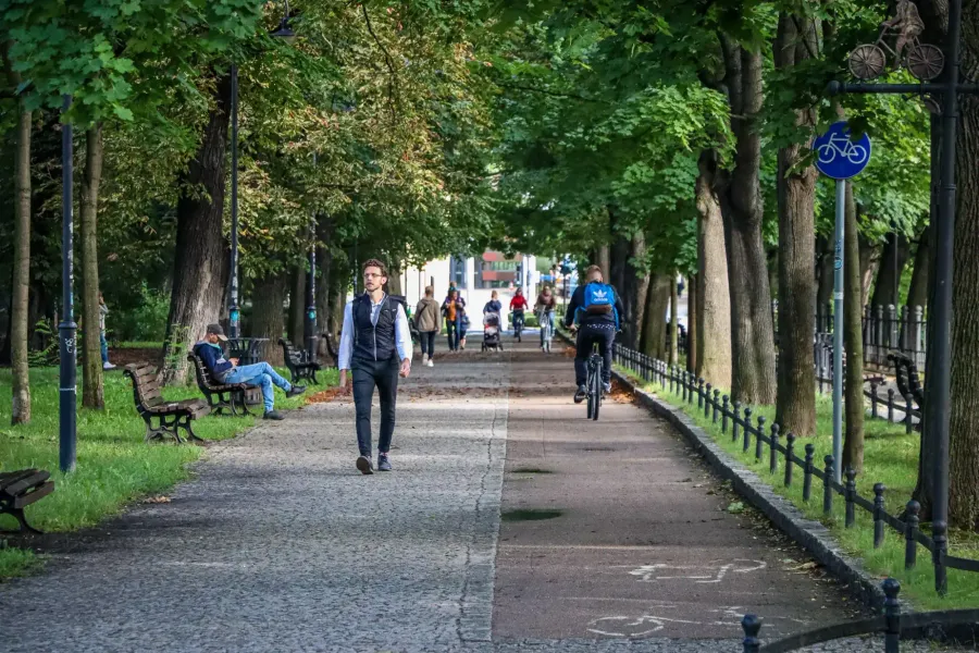 A wide park pathway with a pedestrian section and a bicycle lane, lined with green trees. People are walking, cycling, and...