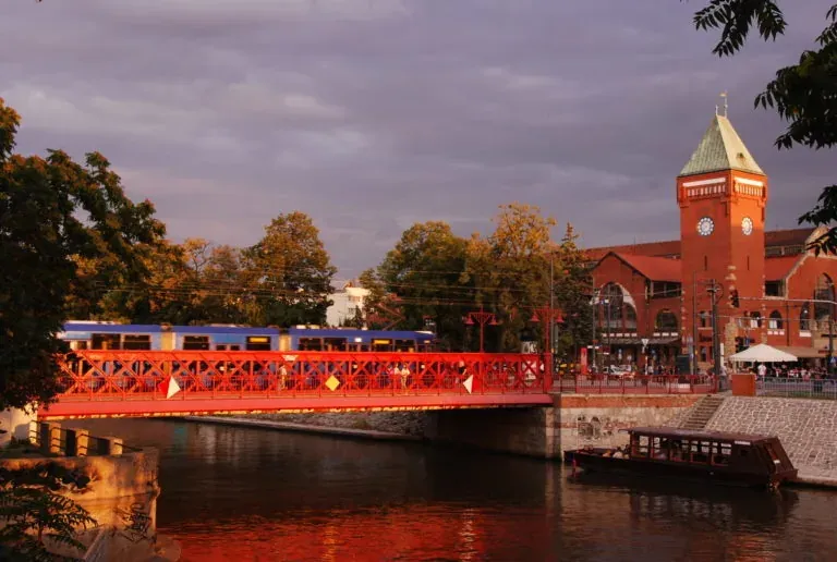 Rote Metallbrücke über einem Fluss, ein blauer Zug fährt darüber, daneben eine rote Backstein-Markthalle mit Uhrturm bei S...