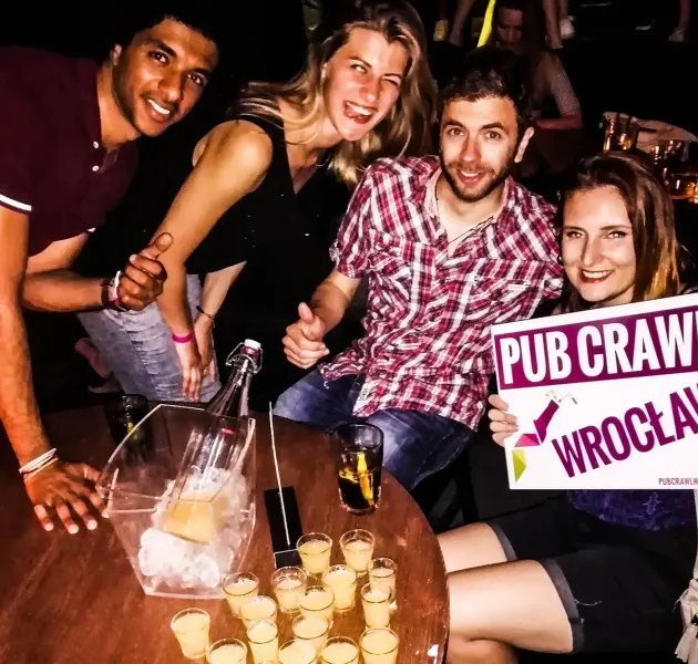 Cztery uśmiechnięte osoby przy stole z napojami i szotami, jedna trzyma znak 'PUB CRAWL WROCŁAW'.